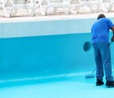 Operaio intento a verniciare una piscina in calcestruzzo con un rullo e una vernice di colore azzurro.
