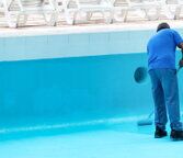 Operaio intento a verniciare una piscina in calcestruzzo con un rullo e una vernice di colore azzurro.